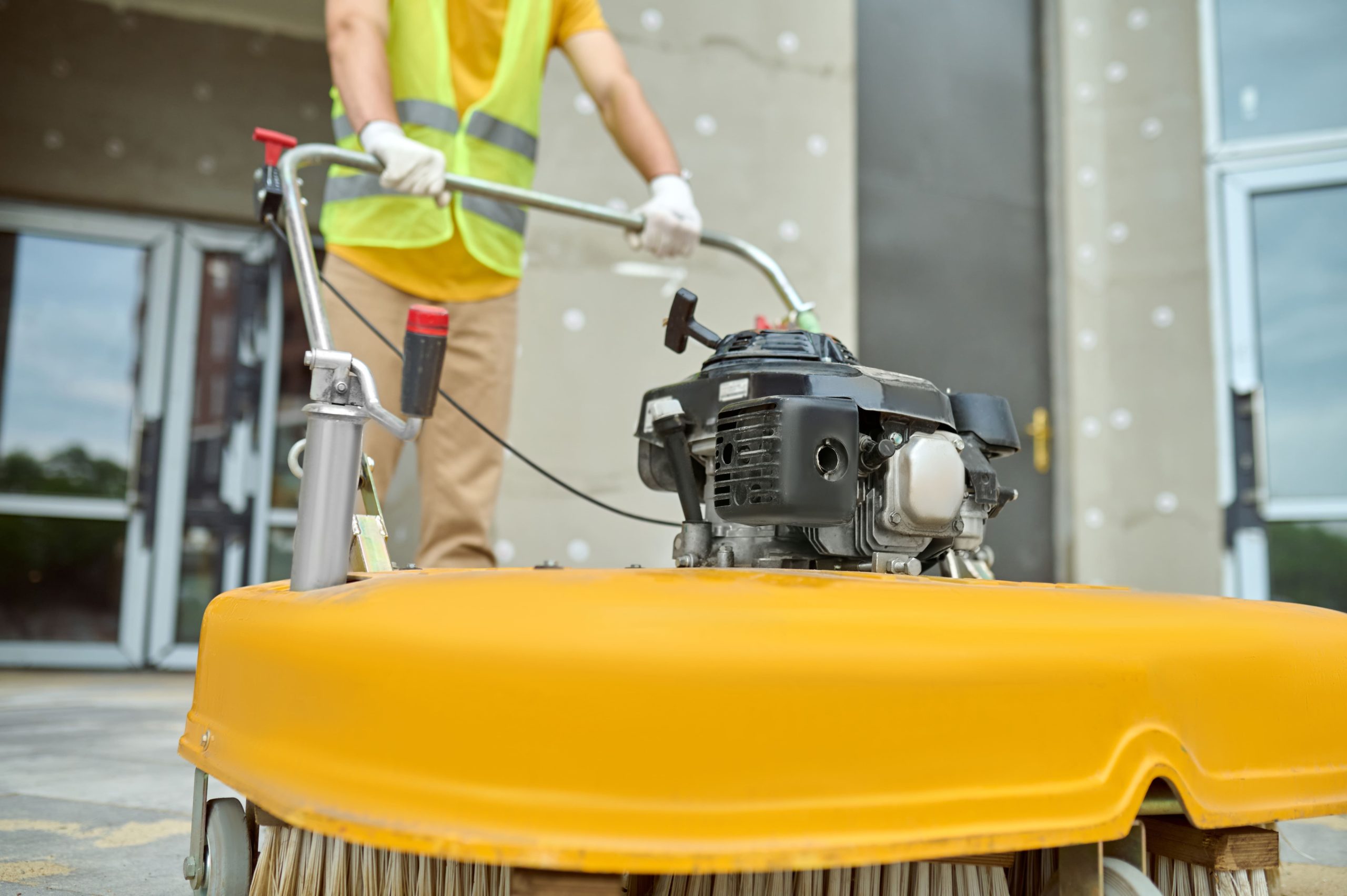 A worker operates a motorized floor sweeper with a yellow brush for outdoor cleaning